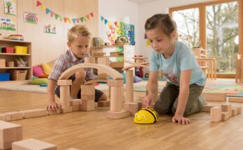 Zwei Kinder spielen in einem hellen Klassenraum mit Holzbausteinen. Ein Junge untersucht eine Baukonstruktion, während ein Mädchen einen programmierbaren Roboter in Form einer Biene steuert. Im Hintergrund sind farbenfrohe Kunstwerke und Spielmaterialien sichtbar, die eine kreative Lernumgebung schaffen.