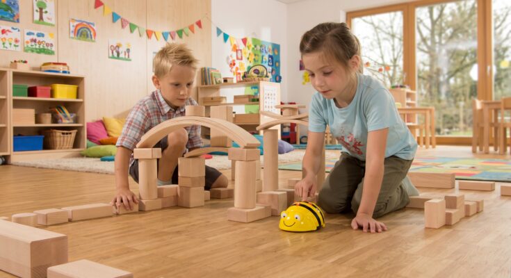 Zwei Kinder spielen in einem hellen Klassenraum mit Holzbausteinen. Ein Junge untersucht eine Baukonstruktion, während ein Mädchen einen programmierbaren Roboter in Form einer Biene steuert. Im Hintergrund sind farbenfrohe Kunstwerke und Spielmaterialien sichtbar, die eine kreative Lernumgebung schaffen.