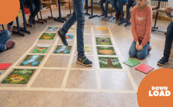 Schüler spielen ein interaktives Lernspiel auf dem Boden, indem sie über große Bilder von Landschaften und Szenen treten. Die Bilder sind auf einem mit Klebeband markierten Raster angeordnet. Im Hintergrund sind weitere Kinder zu sehen, die aufmerksam zuschauen und Materialien in der Hand halten.