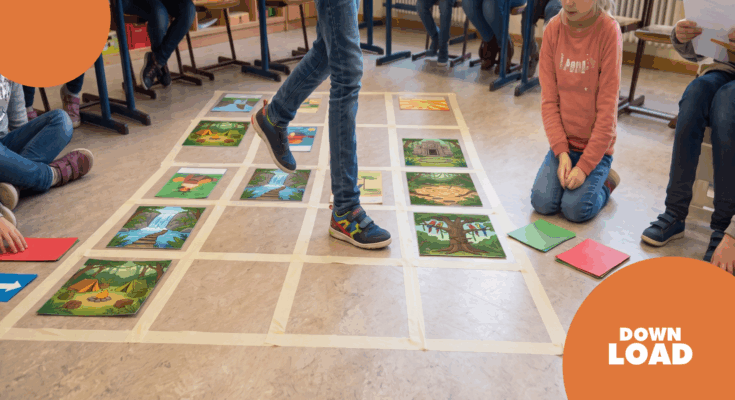 Schüler spielen ein interaktives Lernspiel auf dem Boden, indem sie über große Bilder von Landschaften und Szenen treten. Die Bilder sind auf einem mit Klebeband markierten Raster angeordnet. Im Hintergrund sind weitere Kinder zu sehen, die aufmerksam zuschauen und Materialien in der Hand halten.