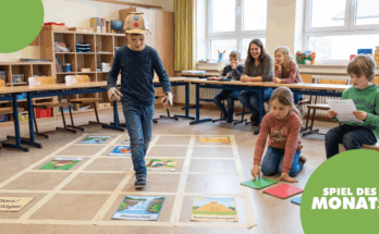 Ein Junge mit einer Pappmütze spielt ein interaktives Lernspiel in einem Klassenzimmer. Um ihn herum sitzen mehrere Kinder und eine Lehrerin, die das Spiel beobachten. Auf dem Boden sind verschiedene Spielkarten mit Motiven wie Wasserfall und Sonnentempel ausgelegt.