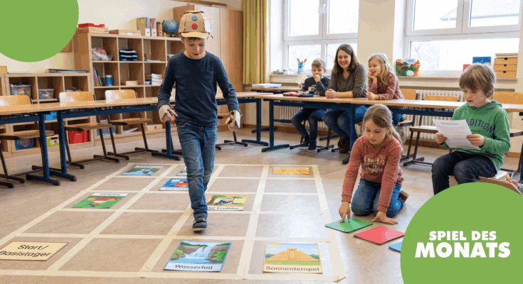 Ein Junge mit einer Pappmütze spielt ein interaktives Lernspiel in einem Klassenzimmer. Um ihn herum sitzen mehrere Kinder und eine Lehrerin, die das Spiel beobachten. Auf dem Boden sind verschiedene Spielkarten mit Motiven wie Wasserfall und Sonnentempel ausgelegt.