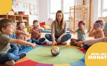 Eine Gruppe von Kindern sitzt im Kreis auf einem bunten Teppich und spielt ein Spiel. Sie tragen Augenbinden und zeigen auf einen Ball, während eine Erwachsene ihnen Anweisungen gibt. Der Raum ist hell und freundlich, mit Spielzeug und bunten Kissen im Hintergrund.