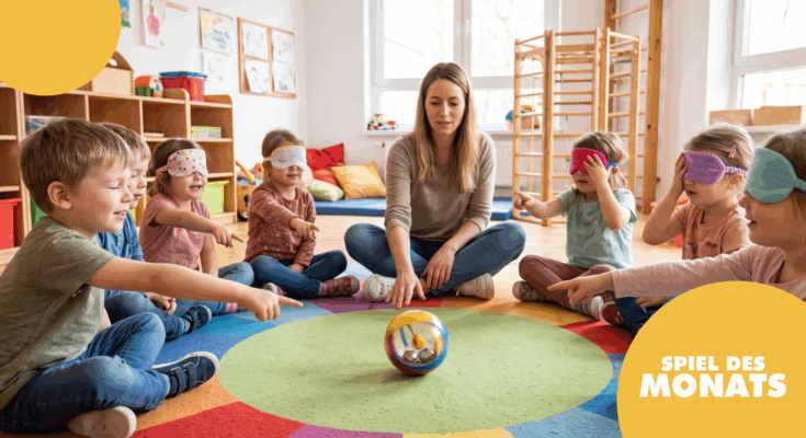 Eine Gruppe von Kindern sitzt im Kreis auf einem bunten Teppich und spielt ein Spiel. Sie tragen Augenbinden und zeigen auf einen Ball, während eine Erwachsene ihnen Anweisungen gibt. Der Raum ist hell und freundlich, mit Spielzeug und bunten Kissen im Hintergrund.