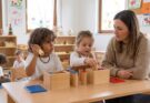 Kinder beim Spielen und Lernen in einem hellen Klassenzimmer. Ein Junge hält ein Spielzeugtelefon, während ein Mädchen mit Holzbausteinen in einer Kiste spielt. Eine Erwachsene beobachtet aufmerksam die Aktivitäten der Kinder. Hintergrund mit bunten Zeichnungen und Lernmaterialien.