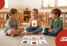 Drei Kinder sitzen auf einem Teppich in einem hellen Raum und spielen mit bunten Karten, die verschiedene Tiere darstellen. Eines der Kinder hält eine Karte mit dem Titel „Sonnen-Fuchs“ hoch und lächelt. Im Hintergrund sind Holzspielzeug und Stühle zu sehen, was eine freundliche Lernumgebung schafft.