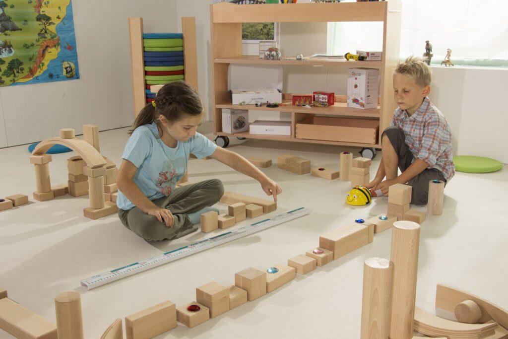 Zwei Kinder spielen in einem hellen Raum mit Holzbausteinen. Ein Mädchen sitzt am Boden und konstruiert eine Struktur, während ein Junge neben ihr mit einem Spielzeugauto spielt. Im Hintergrund sind Regale mit Spielzeug und einer bunten Matratze zu sehen.